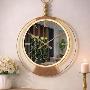 Puede incluir: Reloj de pared dorado con esfera de espejo circular. El reloj tiene un marco dorado con elementos decorativos superiores e inferiores. Las manecillas y los números del reloj son dorados. Una luz cálida ilumina la esfera. Colgando de una pared blanca.