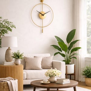 K&ouml;nnte beinhalten: Eine goldfarbene, minimalistische Wanduhr mit schwarzen Zeigern h&auml;ngt an einer wei&szlig;en Wand. Die Uhr hat einen kreisf&ouml;rmigen Rahmen und ein Pendel. Der Raum verf&uuml;gt &uuml;ber ein wei&szlig;es Sofa, einen runden Couchtisch aus Holz und verschiedene Pflanzen.