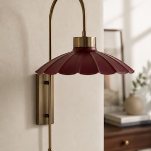 Op de afbeelding: Een wandlamp met een bordeauxrode, geschulpte kap en een messingkleurig metalen frame. De gebogen arm en rechthoekige basis geven een vintage uitstraling. Geschikt voor wandmontage in diverse interieurs.