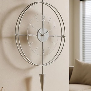 Puede incluir: Un reloj de pared moderno de color plateado con un diseño minimalista. El reloj presenta un marco circular, una esfera de cristal transparente con marcadores de hora simples y un peso de estilo péndulo. El reloj está montado en una pared beige.