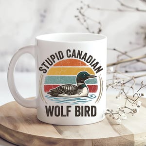 May include: White ceramic mug featuring a retro design with a loon illustration and the text "STUPID CANADIAN WOLF BIRD." The design includes a sunset-style background with orange, yellow, teal, and red stripes. The mug has a curved handle.