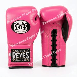 May include: A pair of bright pink boxing gloves with black lace-up closures. The gloves feature the Cleto Reyes logo in black and white ovals, with the text '14oz' and 'Hecho en Mexico' printed on the wrist area of the left glove.