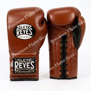 May include: A pair of brown leather boxing gloves with black lace-up closures. The gloves feature the Cleto Reyes logo in a black oval patch on the front and a rectangular label on the wrist displaying the text 14oz and Cleto Reyes Profesional.