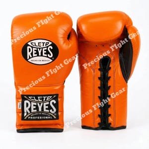 May include: A pair of bright orange boxing gloves featuring black lace-up closures and black trim. The gloves display the Cleto Reyes logo in black and white on the wrist and back of the hand, with the text 14oz clearly visible on the wrist label.