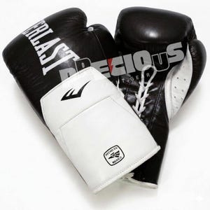 Può includere: Un paio di guanti da boxe neri e bianchi con il logo Everlast in testo bianco sulla superficie nera. I guanti presentano chiusure con lacci bianchi e un polsino bianco con un piccolo emblema nero contenente il testo HECHO EN MEXICO.