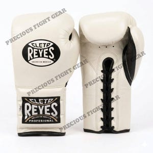 May include: A pair of off-white Cleto Reyes boxing gloves with black lace-up closures and black trim. The gloves feature the Cleto Reyes logo on the wrist and back of the hand, with a label indicating 14oz weight and the text Hecho en Mexico.
