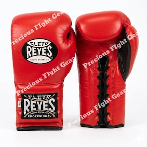 May include: A pair of bright red Cleto Reyes boxing gloves featuring black lace-up closures and black logo patches. The gloves display the text Cleto Reyes Hecho en Mexico and 14oz on the wrist area. The design includes black accents on the palm side.