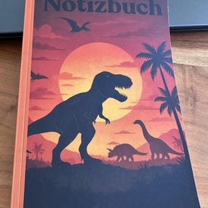 Op de afbeelding: Notitieboek met een dinosaurus-thema omslag. De omslag toont een zonsondergang met silhouetten van dinosaurussen, waaronder een T-Rex. Het woord "Notizbuch" staat bovenaan gedrukt. Het notitieboek heeft een oranje rug.