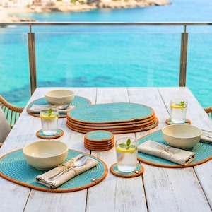May include: A set of oval turquoise woven placemats with orange trim, matching round coasters, beige linen napkins tied with twine, and off-white ceramic bowls arranged on a white wooden table for a complete dining table setting.