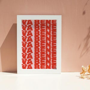 May include: A framed art print with the words "VA BENE" in red, vertically stacked and repeated. The text has a glitch effect. The white frame contrasts with the red text and the pink background.