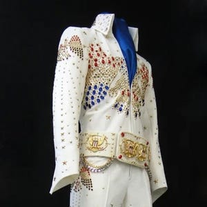 Può includere: Tuta bianca con elaborate decorazioni rosse, blu e oro. La giacca e i pantaloni presentano disegni di stelle e aquile, un'ampia cintura e una sciarpa blu. I pantaloni a zampa hanno dettagli rossi.