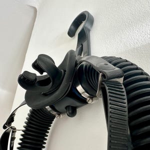 Peut inclure: Masque de plongée sous-marine noir avec un tuyau ondulé et des sangles réglables. Le masque est monté sur un mur blanc, mettant en valeur sa conception et ses composants. Le masque comprend un embout buccal et une sangle sécurisée.