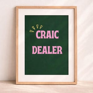 Può includere: Stampa artistica incorniciata con la scritta "CRAIC DEALER" in rosa su sfondo verde scuro, con elementi gialli. La stampa è racchiusa in una cornice di legno chiaro.