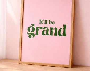 May include: A rectangular wooden frame containing a light pink print with the dark green text It'll be grand centered in the middle. The frame has a natural wood finish and holds a smooth, flat surface with the printed lettering clearly visible.