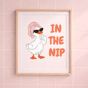 Può includere: Stampa artistica incorniciata con un'oca dei cartoni animati con occhiali da sole e un asciugamano rosa in testa. Il testo "IN THE NIP" è arancione. L'opera è esposta su una parete piastrellata rosa.