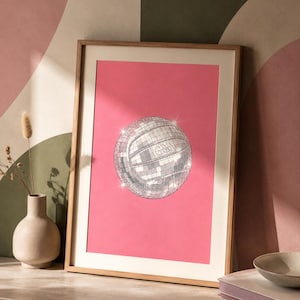 Può includere: Una stampa d'arte verticale incorniciata che presenta un'illustrazione centrale di una pallavolo ricoperta di specchietti argentati riflettenti, creando un effetto palla da discoteca. Lo sfondo della stampa è rosa acceso e la cornice è in legno chiaro.