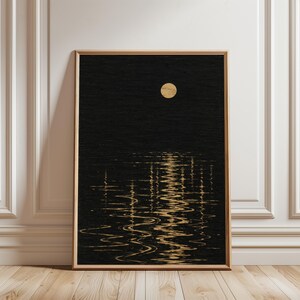 Op de afbeelding: Een rechthoekige wandkunstprint in een lichthouten lijst met een donkere zwarte textuurachtergrond, een enkele gouden cirkelvormige maan in het midden bovenaan en een verticale reflectie van goud licht golvend over de onderste helft.