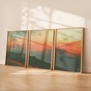 Puede incluir: Tres impresiones artísticas enmarcadas de paisajes. Cada impresión presenta una escena de montaña borrosa con colores verdes, naranjas y amarillos. Los marcos son de color madera clara, y las impresiones se apoyan contra una pared blanca sobre un suelo de madera.