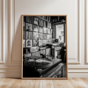 May include: Black and white framed photograph of a vintage record player with a vinyl record on top. The background shows a wall filled with vinyl records. The photograph is displayed on a wooden floor next to a white wall.