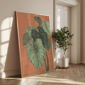 May include: A framed botanical print featuring a detailed illustration of large, green tropical leaves against a muted orange background. The artwork is displayed in a room with a wooden floor and a small potted plant.