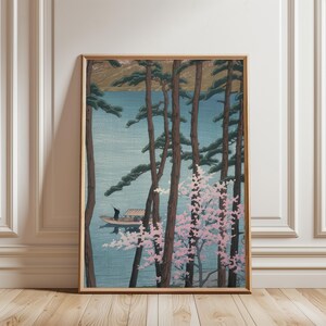 May include: Framed art print depicting a serene lake scene. Tall, slender trees frame the view of a boat on the water. Pink flowering branches add a touch of color against the blue water and sky. The artwork is in a wooden frame.