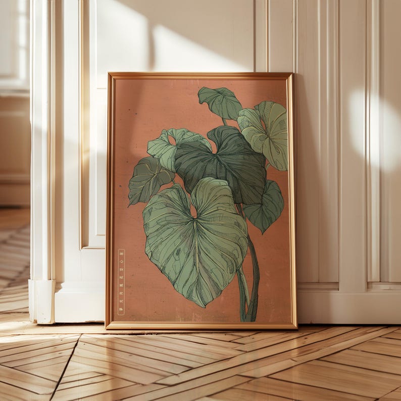 May include: A framed botanical print featuring a detailed illustration of large, green leaves against a peach-colored background. The artwork is enclosed in a gold-colored frame, adding a touch of elegance. The print is leaning against a white door.