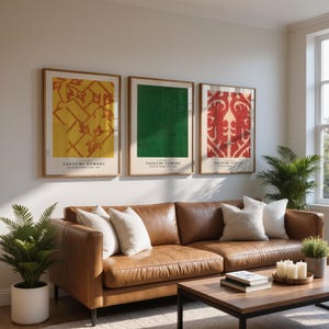 Puede incluir: Tres impresiones artísticas enmarcadas con diseños abstractos en amarillo, verde y rojo cuelgan sobre un sofá de cuero marrón. Las impresiones presentan el texto "TAGUCHI TOMOKI" y se exhiben en un salón bien iluminado con plantas y una mesa de centro.