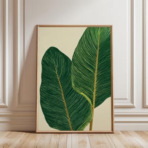 May include: A framed art print depicting two large, stylized green leaves with detailed vein patterns. The leaves have a yellow-gold stem against a cream background. The frame is a light brown colour.