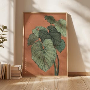May include: A framed botanical print featuring a detailed illustration of large, green leaves against a muted orange background. The artwork is in a wooden frame and includes the text "MODERN MUSEUM" vertically on the side. The print is displayed on a wooden floor.