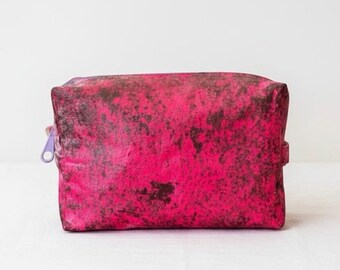 Puede incluir: Un neceser rectangular de color rosa intenso con un aspecto texturizado y moteado. La bolsa tiene una cremallera morada y una pequeña correa lateral. La bolsa está sobre una superficie blanca.