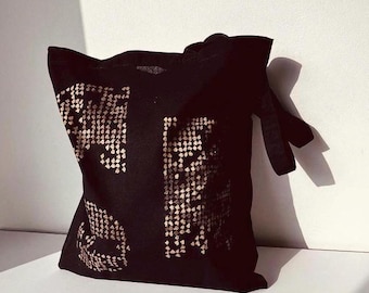 Puede incluir: Un bolso tote negro con un gran estampado gráfico en color dorado metálico con patrón de piel de serpiente en la parte frontal. El bolso tiene dos asas de tela negra y una forma rectangular, con el diseño dorado formando dos secciones.
