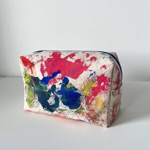 Puede incluir: Un neceser rectangular de lona blanquecina con cremallera azul. La bolsa est&aacute; decorada con vibrantes salpicaduras de pintura roja, azul, amarilla y verde. Dise&ntilde;ado para guardar maquillaje y otros art&iacute;culos personales.