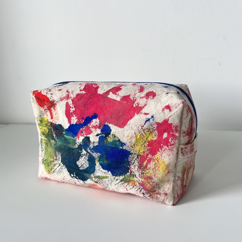 Puede incluir: Un neceser rectangular de lona blanquecina con cremallera azul. La bolsa est&aacute; decorada con vibrantes salpicaduras de pintura roja, azul, amarilla y verde. Dise&ntilde;ado para guardar maquillaje y otros art&iacute;culos personales.