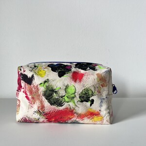 Puede incluir: Un neceser rectangular con un fondo blanco cubierto de pinceladas coloridas. Las pinceladas son de varios colores, incluyendo rojo, verde, amarillo y negro. La bolsa tiene una cremallera azul.