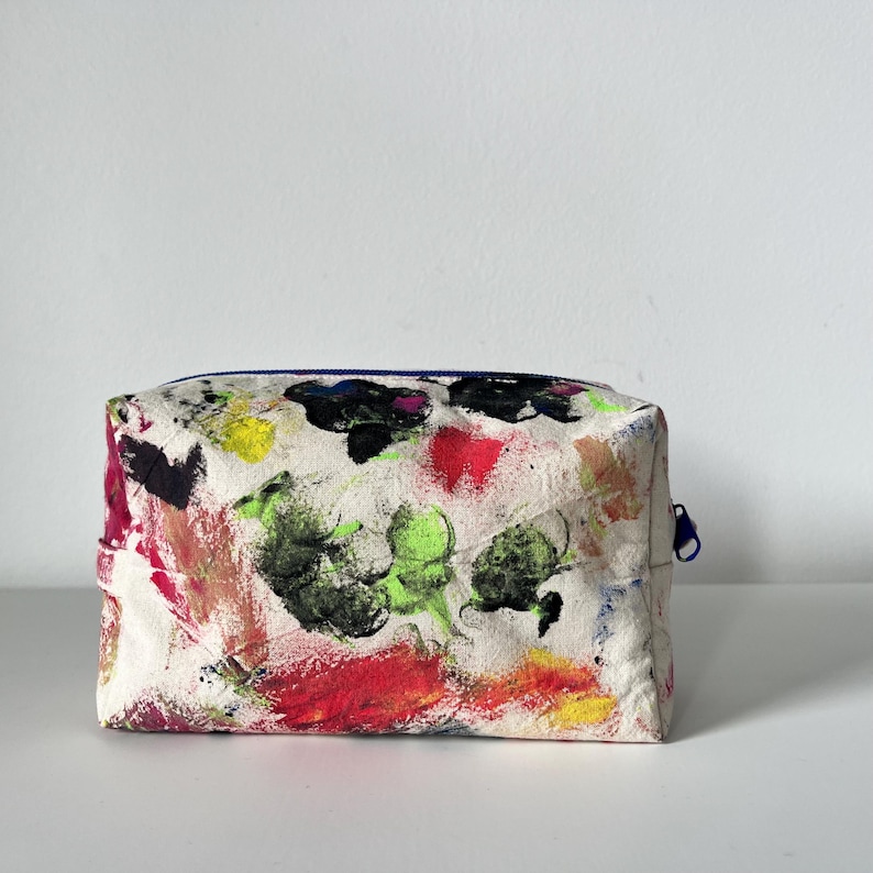 Puede incluir: Un neceser rectangular con un fondo blanco cubierto de pinceladas coloridas. Las pinceladas son de varios colores, incluyendo rojo, verde, amarillo y negro. La bolsa tiene una cremallera azul.