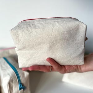 Puede incluir: Una bolsa rectangular de color crema con un forro de cremallera rojo se sostiene en una mano. La bolsa parece estar hecha de una tela texturizada. Otras bolsas con cremalleras de diferentes colores son visibles en el fondo.