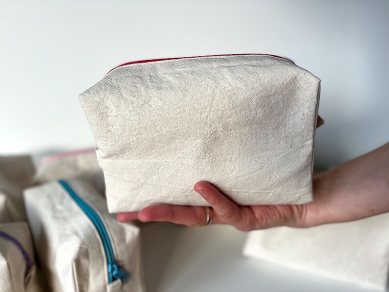 Puede incluir: Una bolsa rectangular de color crema con un forro de cremallera rojo se sostiene en una mano. La bolsa parece estar hecha de una tela texturizada. Otras bolsas con cremalleras de diferentes colores son visibles en el fondo.