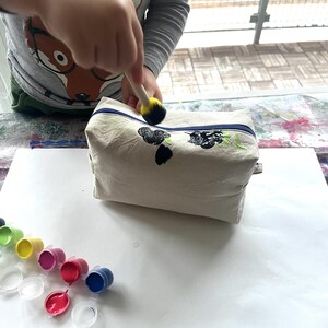 Puede incluir: Un neceser de lona beige con cremallera azul est&aacute; siendo pintado con pintura negra y verde. Varios peque&ntilde;os recipientes de pintura de varios colores son visibles sobre la mesa. Un ni&ntilde;o est&aacute; usando un pincel de esponja para aplicar la pintura.