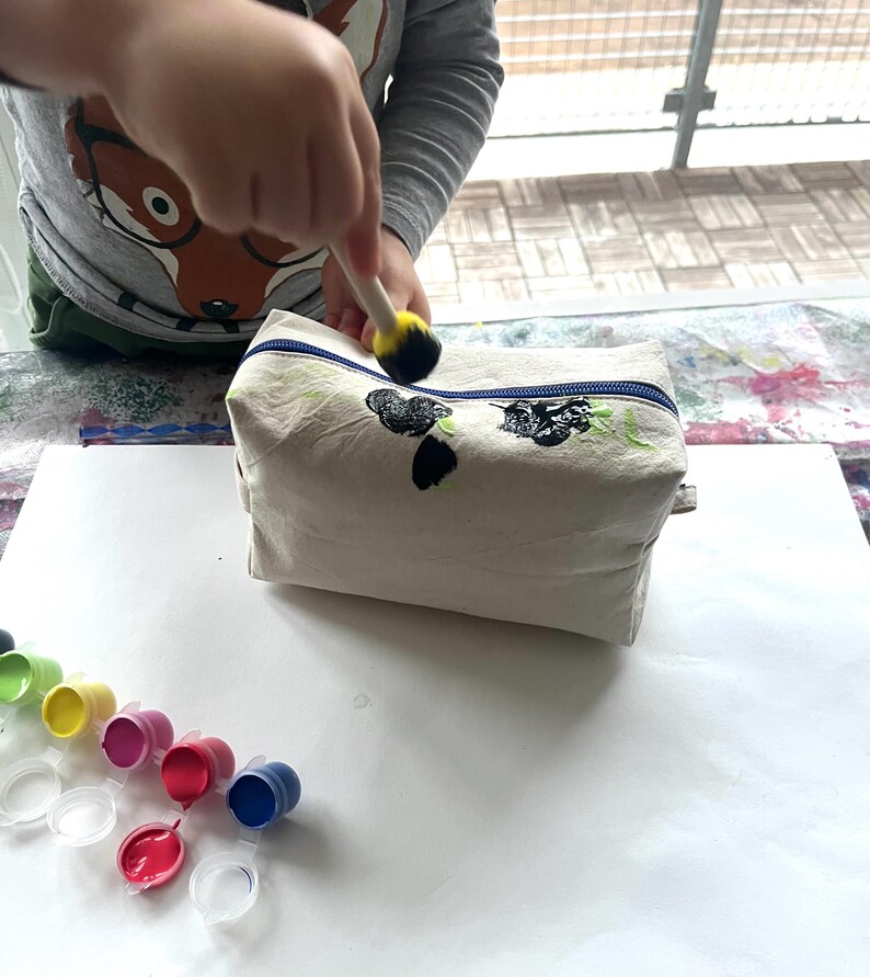 Puede incluir: Un neceser de lona beige con cremallera azul est&aacute; siendo pintado con pintura negra y verde. Varios peque&ntilde;os recipientes de pintura de varios colores son visibles sobre la mesa. Un ni&ntilde;o est&aacute; usando un pincel de esponja para aplicar la pintura.