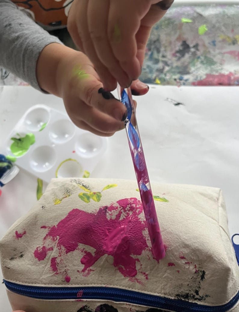 Puede incluir: Un estuche de lona beige con cremallera azul est&aacute; salpicado de pintura rosa. Un pincel con pintura rosa y azul se sostiene sobre el estuche. Una paleta de pintura blanca con pintura verde est&aacute; en el fondo.