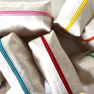 Puede incluir: Un conjunto de neceseres rectangulares beige con cremalleras de colores. Las cremalleras son de varios colores, incluyendo rosa, amarillo, rojo, naranja, verde y turquesa. Los neceseres est&aacute;n hechos de un material texturizado.