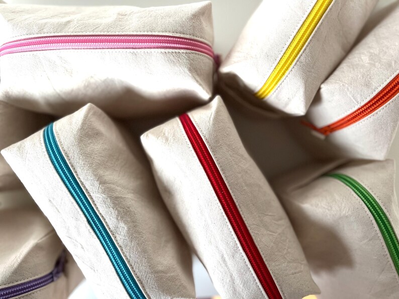 Puede incluir: Un conjunto de neceseres rectangulares beige con cremalleras de colores. Las cremalleras son de varios colores, incluyendo rosa, amarillo, rojo, naranja, verde y turquesa. Los neceseres est&aacute;n hechos de un material texturizado.