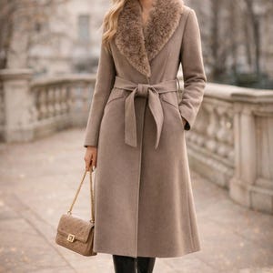 Peut inclure: Long manteau taupe avec col en fausse fourrure et ceinture assortie. Le manteau pr&eacute;sente un design classique avec un ourlet au genou et est associ&eacute; &agrave; des bottes noires et un sac &agrave; main beige avec une cha&icirc;ne dor&eacute;e. La longueur du manteau est d'environ 120 cm.