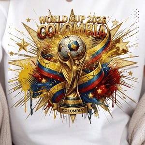 May include: A white short-sleeved t-shirt featuring a graphic print of the World Cup 2026 Colombia logo. The design includes a golden trophy, a soccer ball, and ribbons in yellow, blue, and red colors, surrounded by golden starbursts.