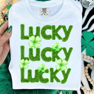 Puede incluir: Camiseta blanca con la palabra "LUCKY" en letras grandes, verdes y texturizadas, cada una con un trébol. La camiseta tiene cuello redondo y una etiqueta que dice "COMFORT COLORS". Un diseño festivo del Día de San Patricio.