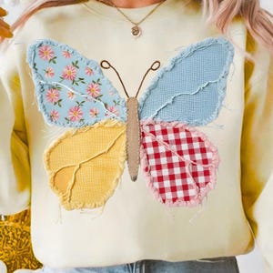 May include: A cream-colored sweatshirt features a large, patchwork butterfly design. The butterfly's wings are made of various fabrics, including floral, gingham, and solid colors. The butterfly has brown antennae and a tan body.