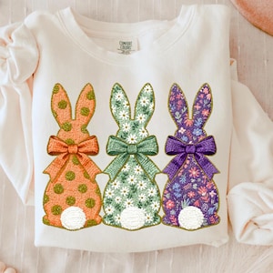 Könnte beinhalten: Cremefarbenes Sweatshirt mit drei gestickten Hasenmotiven. Die Hasen haben gemusterte Körper in Orange, Grün und Lila, jeweils mit passender Schleife. Ideal für Ostern oder den Frühling.