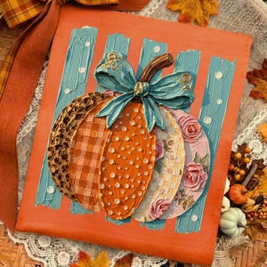 May include: A square orange canvas featuring a textured pumpkin painting with a blue bow on top. The pumpkin segments display varied patterns including leopard print, orange plaid, white polka dots, and pink roses on a light blue striped background.