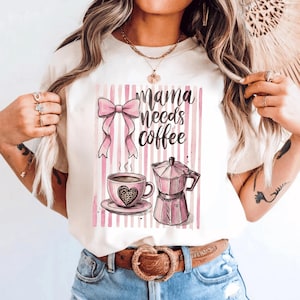 Könnte beinhalten: Cremefarbenes T-Shirt mit rosa Schleife, dem Schriftzug "Mama needs coffee", einer Kaffeetasse mit Herzmuster und einer rosa Kaffeemaschine. Der Hintergrund hat rosa und weiße vertikale Streifen.