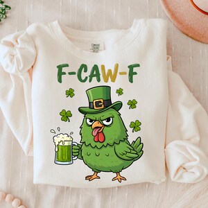 Op de afbeelding: Crèmekleurige sweatshirt met een groene afbeelding van een kip met een leprechaun-hoed en een groen bier. De tekst "F-CAW-F" staat boven de kip, met klavers rond het ontwerp. De sweatshirt is gemaakt van zacht materiaal.